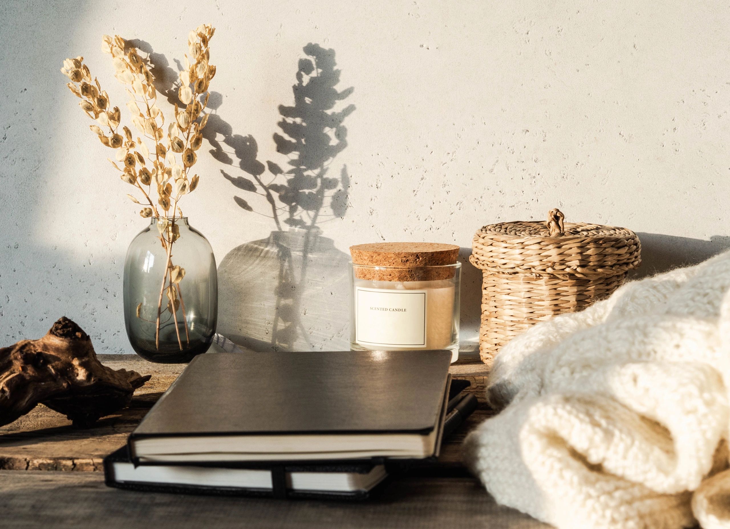 Cozy autumn home still life of dry flowers in vase, straw box, glass candle, knitted blanket and black notepads on a rustic wooden table and shadows on the wall from the bright setting sun